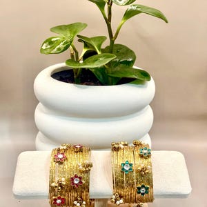 May include: Two gold bangles with floral and pearl accents rest on a white display stand. The bangles feature red and green gemstones. A white, layered ceramic planter with a green plant sits in the background.