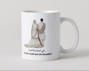 Op de afbeelding: Witte keramische mok met een huwelijksthema-illustratie. Het toont een stel in traditionele kledij, met de bruid in een witte jurk. De mok bevat Arabische en Engelse tekst: 'In this world and the hereafter'.