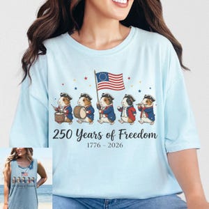 May include: Light blue t-shirt featuring a patriotic design. The graphic includes guinea pigs dressed in colonial attire, marching with a flag. Text reads "250 Years of Freedom 1776-2026". Stars and stripes complete the design.