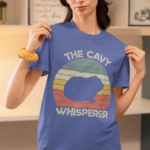 May include: A blue t-shirt with a retro design. It features a guinea pig silhouette inside a striped circle. The text reads "THE CAVY WHISPERER" in a vintage font. The shirt is a solid blue colour.