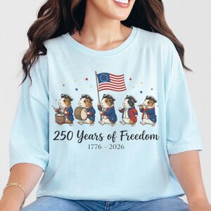 May include: Light blue t-shirt featuring a patriotic design. The graphic shows five guinea pigs dressed in colonial attire, marching with a flag. Text reads "250 Years of Freedom 1776-2026".