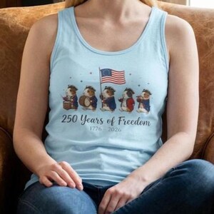 May include: Light blue tank top featuring a design with guinea pigs dressed in colonial attire, holding a flag and musical instruments. The text "250 Years of Freedom 1776-2026" is printed below the illustration. The tank top is a casual style.
