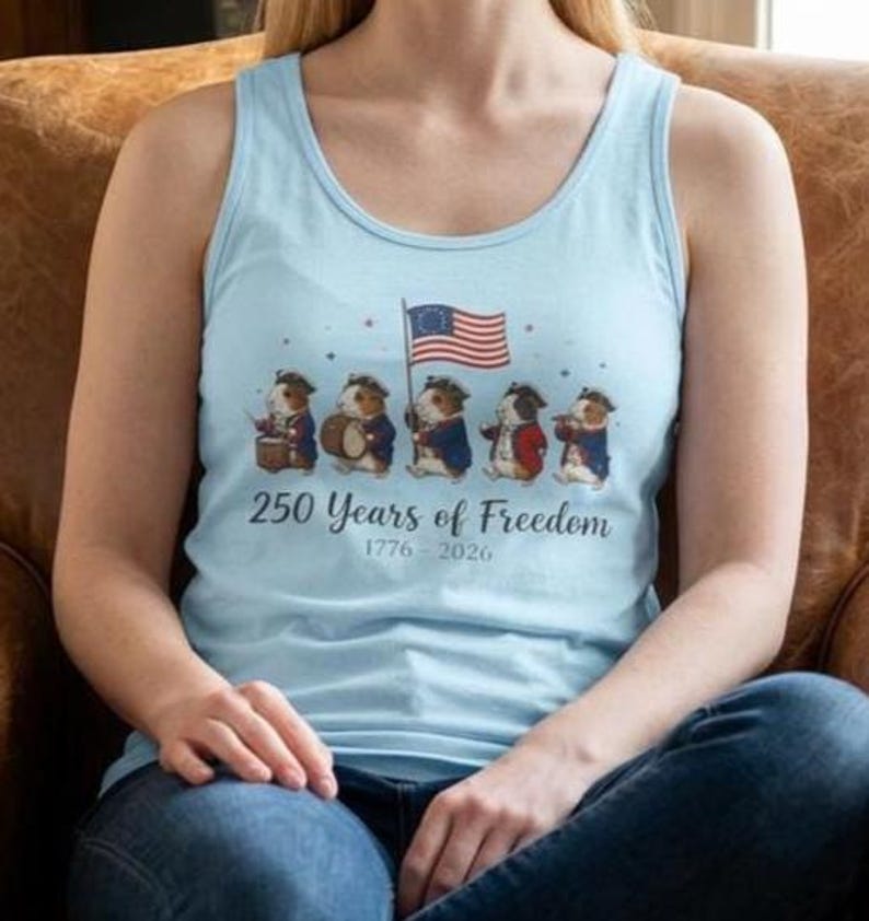 May include: Light blue tank top featuring a design with guinea pigs dressed in colonial attire, holding a flag and musical instruments. The text "250 Years of Freedom 1776-2026" is printed below the illustration. The tank top is a casual style.