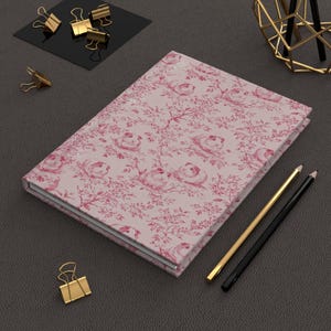 May include: A closed journal with a pink floral design of roses and birds. The cover is hardback. Two pencils, one gold and one black, are placed beside the journal. Gold binder clips are also visible.