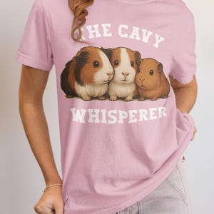 May include: A pale pink t-shirt with the words "THE CAVY WHISPERER" above a graphic of three guinea pigs. The guinea pigs are brown, white, and tan. The t-shirt has a crew neck and short sleeves.