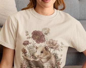 May include: Cream-colored t-shirt featuring a detailed illustration of a hamster reading a book amidst flowers. The design includes shades of brown, pink, and cream, creating a vintage aesthetic. The shirt is a classic crew neck style.
