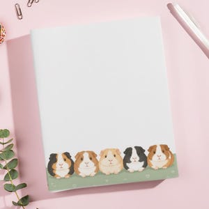 May include: A white notepad with a decorative border featuring five guinea pig illustrations in various colors. The notepad is surrounded by stationery items, including washi tape, paper clips, and a pen, all set against a pink background.