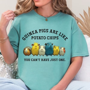 Puede incluir: Una camiseta de color turquesa con el texto "GUINEA PIGS ARE LIKE POTATO CHIPS YOU CAN'T HAVE JUST ONE." La camiseta presenta un gráfico de seis cobayas en varios colores, incluyendo amarillo, azul y blanco.