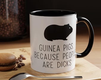 May include: White ceramic mug with a black interior and handle. The mug features a black silhouette of a guinea pig and the text "GUINEA PIGS BECAUSE PEOPLE ARE DICKS" in black. A spoon and a macaron are on a wooden surface.