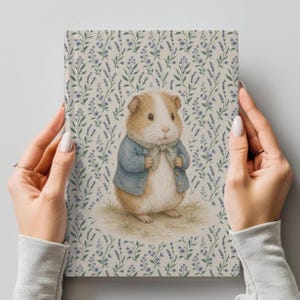 Puede incluir: Un diario de tapa dura con una ilustración caprichosa de un conejillo de indias con una chaqueta azul y pajarita. La portada tiene un estampado floral en tonos morados y verdes sobre un fondo crema. El diario está sostenido por dos manos.