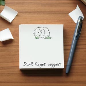 Op de afbeelding: Een vierkant wit notitieblok met een zwarte lijntekening van een cavia die bovenaan een groen bladgroente eet en de zwarte handgeschreven tekst Don't forget veggies! onderaan, vergezeld van een grijze balpen.
