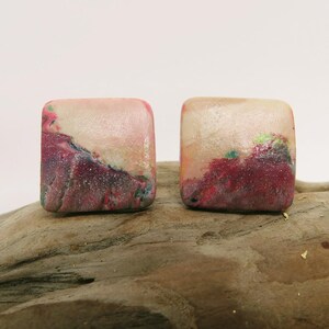 May include: Two square earrings with a marbled effect. The earrings have a blend of pink, red, blue, and green hues, with a lighter top section. They are presented on a piece of driftwood, showcasing their unique design.