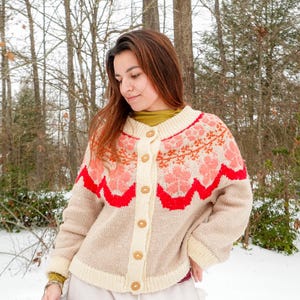 May include: A beige, button-down cardigan sweater with a floral pattern in shades of pink and orange. The sweater has a red scalloped trim and wooden buttons. The background shows a snowy outdoor scene with trees.