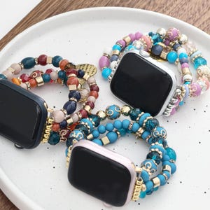May include: Three beaded watch bands in various colors, including blue, pink, and gold, are displayed on a white plate. The bands feature a mix of beads and decorative elements, creating a stylish accessory. The watch faces are black.