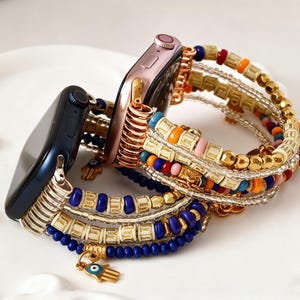 Puede incluir: Dos smartwatches con correas decorativas. Uno es azul marino, el otro es oro rosa. Las correas presentan cuentas doradas, azules, naranjas y blancas, con un dije dorado de una mano con un ojo.