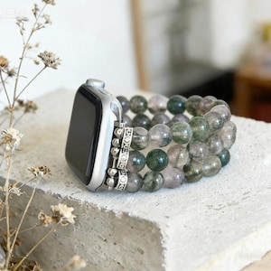 May include: A silver smartwatch case attached to a triple-strand bracelet made of round, translucent green and grey moss agate beads. The bracelet features small, decorative silver-toned metal spacers between the bead strands and the watch.