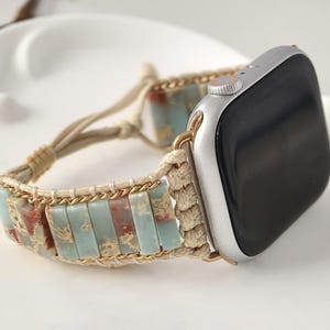 Puede incluir: Un reloj inteligente plateado con pantalla negra está sujeto a una correa de reloj hecha a mano. La correa presenta cuentas rectangulares multicolores en tonos azules, rojos y beige, conectadas por una cadena dorada y una cuerda beige.