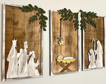 Peut inclure: Ensemble de trois panneaux muraux en bois représentant une scène de la Nativité. Chaque panneau présente un fond en bois brun avec des personnages blancs, des accents de feuilles vertes et un cadre blanc. Les panneaux montrent les Rois Mages, la Nativité et la Sainte Famille.