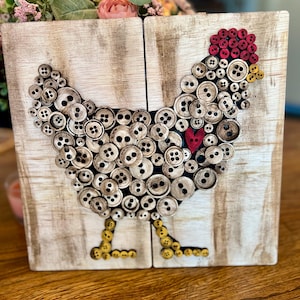 May include: A decorative wooden block featuring a chicken design made from various buttons. The chicken's body is composed of beige buttons, with a red button comb and a red heart. The legs are yellow buttons. The background is a light wood grain.