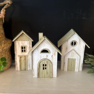 May include: Three miniature wooden houses with white and light green paint. The houses have varying architectural details, including arched doorways, windows, and chimneys. The houses are arranged on a white surface, with a woven basket and greenery in the background.