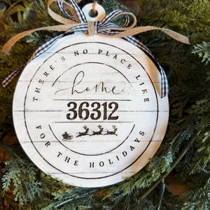 May include: A round white wooden ornament featuring the black text "THERE'S NO PLACE LIKE home 36312 FOR THE HOLIDAYS" and a graphic of a sleigh with reindeer. It is topped with a black and white gingham ribbon and a jute twine bow.
