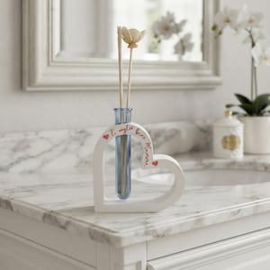 May include: A white heart-shaped wooden frame holding a blue glass test tube vase. Inside the vase are two wooden reed diffuser sticks with a flower topper. The frame features the red text Ti voglio bene Mamma and two small red heart icons.