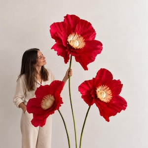 May include: Three oversized, realistic red poppy flowers with textured petals and cream-colored centers, each mounted on a tall, slender green stem. The stems are attached to a flat, square green base, creating a large-scale floral sculpture.