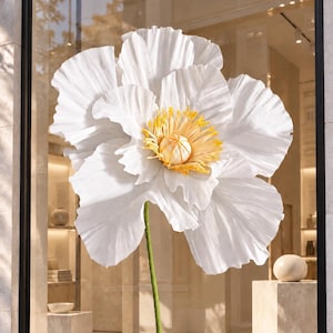May include: A large, oversized decorative flower sculpture featuring expansive, crinkled white petals and a prominent, textured yellow and white center. The flower is supported by a slender green stem attached to a flat, square green metal base.