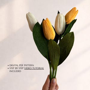 Puede incluir: Un ramo de tulipanes de papel en blanco y amarillo, con hojas verdes. Las flores se sostienen en una mano. El texto en la imagen dice: DIGITAL PDF PATTERN, STEP BY STEP VIDEO TUTORIAL INCLUDED.