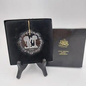 May include: A clear lead crystal ornament featuring an etched figure placing mail in a mailbox with the year 1991 inscribed below. The ornament is displayed in a black box labeled The Enesco Precious Moments Collection Lead Crystal Dated Ornament.