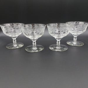 May include: Four clear champagne coupe glasses with etched floral designs. The glasses have a rounded bowl, a short stem, and a circular base. The etched design is visible on the bowl of each glass. The glasses are arranged on a dark surface.