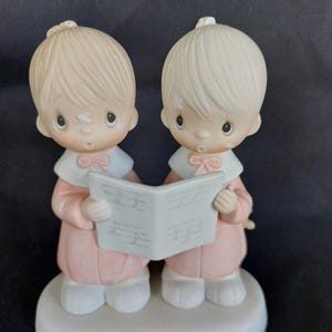 May include: A ceramic figurine depicting two children singing from a music sheet. The figures wear pink robes with white collars and matching bows. The figurine is on a white base, with a dark background.