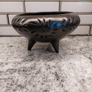 May include: A black, three-legged ceramic bowl with decorative cut-out leaf and dot patterns around the rim. The bowl has a rounded shape and sits on a speckled countertop against a white tile backsplash.