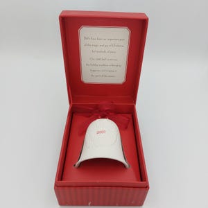 May include: A white ceramic bell ornament with the year 2007 printed in red, topped with a red ribbon. It is presented in an open red box featuring a printed message about the history of Christmas bells on the inside lid.
