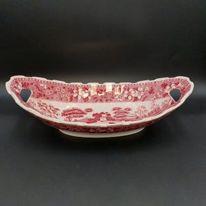 May include: An oval ceramic serving dish featuring a red and white transferware pattern of floral motifs and a landscape scene. The dish has a scalloped rim and two integrated handles on the sides, with a white interior and a red patterned exterior.