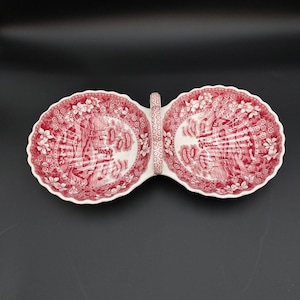 May include: A double-sectioned ceramic serving dish featuring a red and white transferware pattern of floral borders and landscape scenes. The dish has scalloped edges and a central vertical handle with a matching red and white floral design.