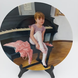 May include: A round decorative plate featuring a painting of a young person with light brown hair wearing a lavender leotard and white tights, sitting on a black piano bench while putting on pink ballet slippers, with a black piano in the background.