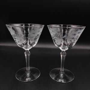 May include: Two clear, stemmed cocktail glasses with etched floral designs. The glasses have a wide, conical bowl, a slender stem, and a circular base. The floral pattern is delicate and wraps around the bowl.