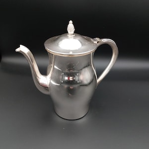 May include: A polished silver-toned metal teapot featuring a curved spout, a sturdy handle, and a removable lid topped with a small, textured finial shaped like a pineapple. The surface of the teapot is smooth and reflective with a metallic finish.