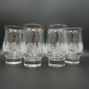 May include: A set of four clear glass tumblers with a curved silhouette and a gold-tone rim. Each glass features a frosted white etched design depicting a winter forest scene with snow-covered pine trees and falling snowflakes around the exterior.