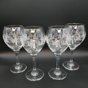 May include: A set of four clear glass wine glasses featuring a delicate etched winter forest scene with pine trees and snow. Each glass has a thin gold rim around the top edge, a rounded bowl, a slender stem, and a circular base.