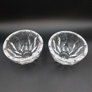 May include: Two clear glass bowls featuring a faceted, starburst-like pattern on the exterior. Each bowl has a circular rim and a deep, rounded interior with reflective surfaces that catch the light, creating a bright and transparent appearance.