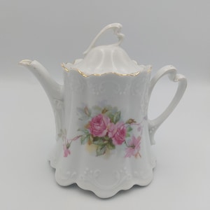 May include: A white porcelain teapot featuring a scalloped base and rim with gold trim. The body is decorated with a pink rose floral pattern and embossed scrollwork. It includes a matching lid with a heart-shaped handle and a curved spout.