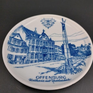 May include: A round white ceramic decorative plate featuring a blue ink illustration of the Offenburg town hall and the Ursulasäule column. The text "OFFENBURG Rathaus mit Ursulasäule" is printed in blue at the bottom, with a small crest above.