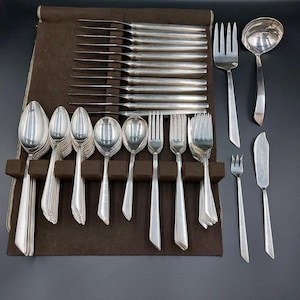 May include: A complete set of silver-colored flatware, including knives, forks, spoons, and a ladle, arranged in a brown felt-lined case. The set includes various sizes of forks and spoons, butter knives, and a serving spoon.