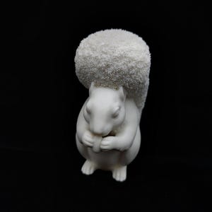 May include: A white squirrel figurine with a smooth matte body and a textured, fuzzy, rounded tail. The squirrel is depicted in a seated position, holding a small object between its front paws, with its large, bushy tail arched over its back.