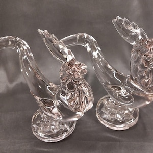 May include: Two clear glass rooster figurines. Each rooster has a detailed head, a curved neck, and a round base. The glass is transparent, allowing light to pass through, and the roosters are positioned on a gray surface.