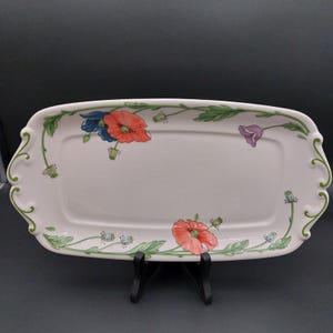 May include: A rectangular white ceramic serving platter with scalloped edges and green scrollwork accents. The surface features a floral pattern with orange-red poppies, a single purple flower, and small blue buds connected by green stems and leaves.