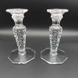 May include: A pair of clear glass candle holders featuring a textured, diamond-patterned stem and a wide, hexagonal base. Each holder has a flared top rim designed to support a candle, with a transparent finish that reflects light across the surface.