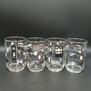 May include: A set of four clear glass tumblers featuring a repeating black and gold printed pattern of interior home furnishings, including a grandfather clock, a hutch, a globe, a rocking chair, and various framed wall art pieces.